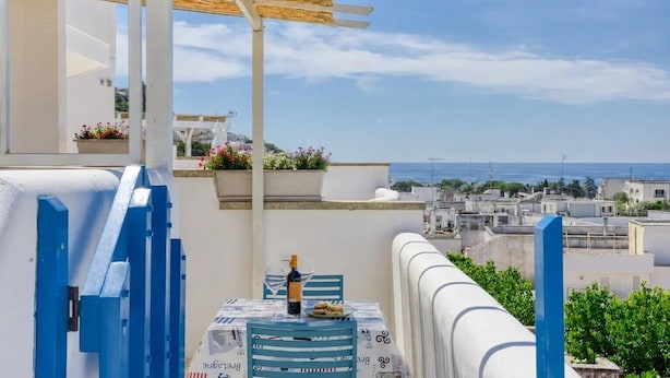 Ein Balkon mit Meerblick, ein Tisch mit Flasche und Gläsern sowie ein blauer Geländer.