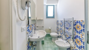 Une salle de bain comprenant un lavabo, des toilettes et un bidet, dont le carrelage présente des motifs bleus et jaunes, et une fenêtre située au-dessus des toilettes.