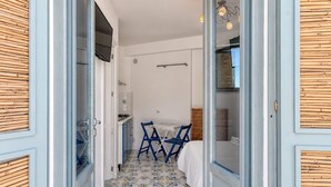 Un couloir étroit avec un coin cuisine, une table à manger entourée de chaises bleues et un lit avec une literie blanche.