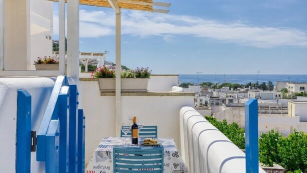 Un balcon avec vue sur la mer, une table avec une bouteille et des verres, et une rambarde bleue.