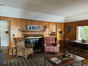 A cozy living room with a fireplace, a plaid armchair, a wicker chair, a coffee table, and a dining area with chairs and a table. - Cozy Lakefront Cottage on Pleasant Lake's Lakeshore Drive (New London)