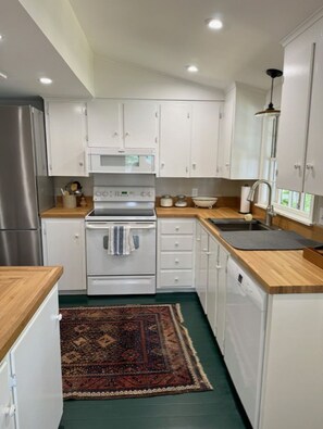A compact kitchen with white cabinets, a stainless steel refrigerator, a microwave, an oven, a sink, and a wooden countertop. - Cozy Lakefront Cottage on Pleasant Lake's Lakeshore Drive (New London)