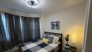 Une chambre à coucher avec un lit, un ventilateur de plafond, une table de chevet avec une lampe, un tableau encadré au mur et une fenêtre avec des rideaux.