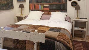 Ein Schlafzimmer mit einem großen Bücherregal, einem Bett mit einer Steppdecke, einem Nachttisch und einem Tisch mit einer Blumen Tischdecke.