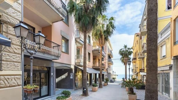 Una calle estrecha flanqueada por edificios de varios pisos, algunos con balcones, y macetas con plantas a lo largo de la acera.