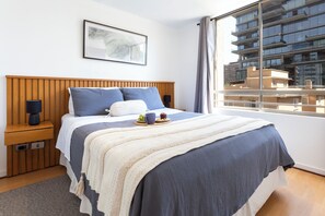 Ein modernes Hotelzimmer mit einem Holzkopfstück, einem großen Fenster mit Blick auf die Stadt, einem Nachttisch mit einer Lampe und einem gerahmten Kunstwerk an der Wand.