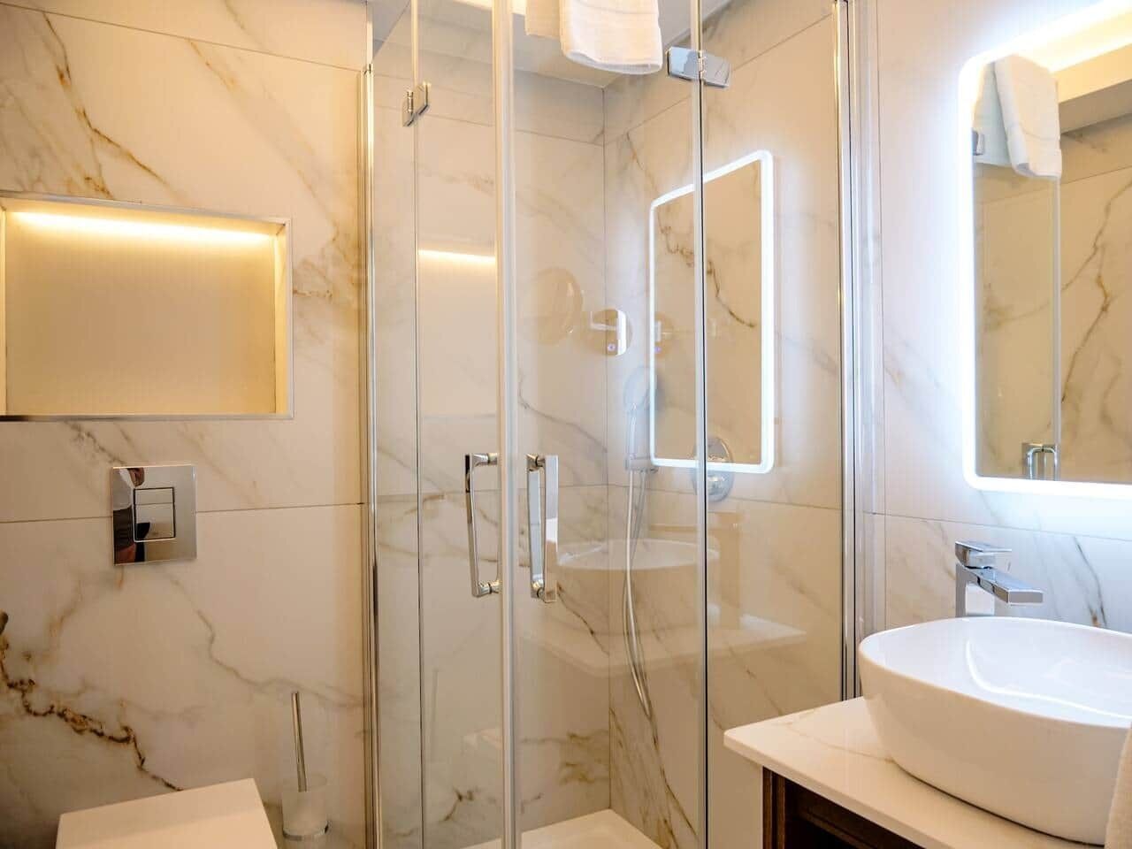 Un baño moderno con ducha de vidrio, pared con azulejos de mármol, lavamanos blanco y un espejo.