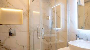 Un baño moderno con ducha de vidrio, pared con azulejos de mármol, lavamanos blanco y un espejo.