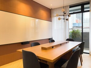 Un salón de conferencias con una larga mesa, sillas, una pantalla de proyector y vistas de edificios a través de amplios ventanales.