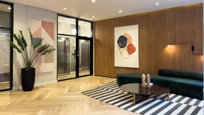 A modern lobby with a herringbone wooden floor, a green sofa, a glass coffee table, a potted plant, and a large abstract painting on the wall. - Today Butanã (São Paulo)