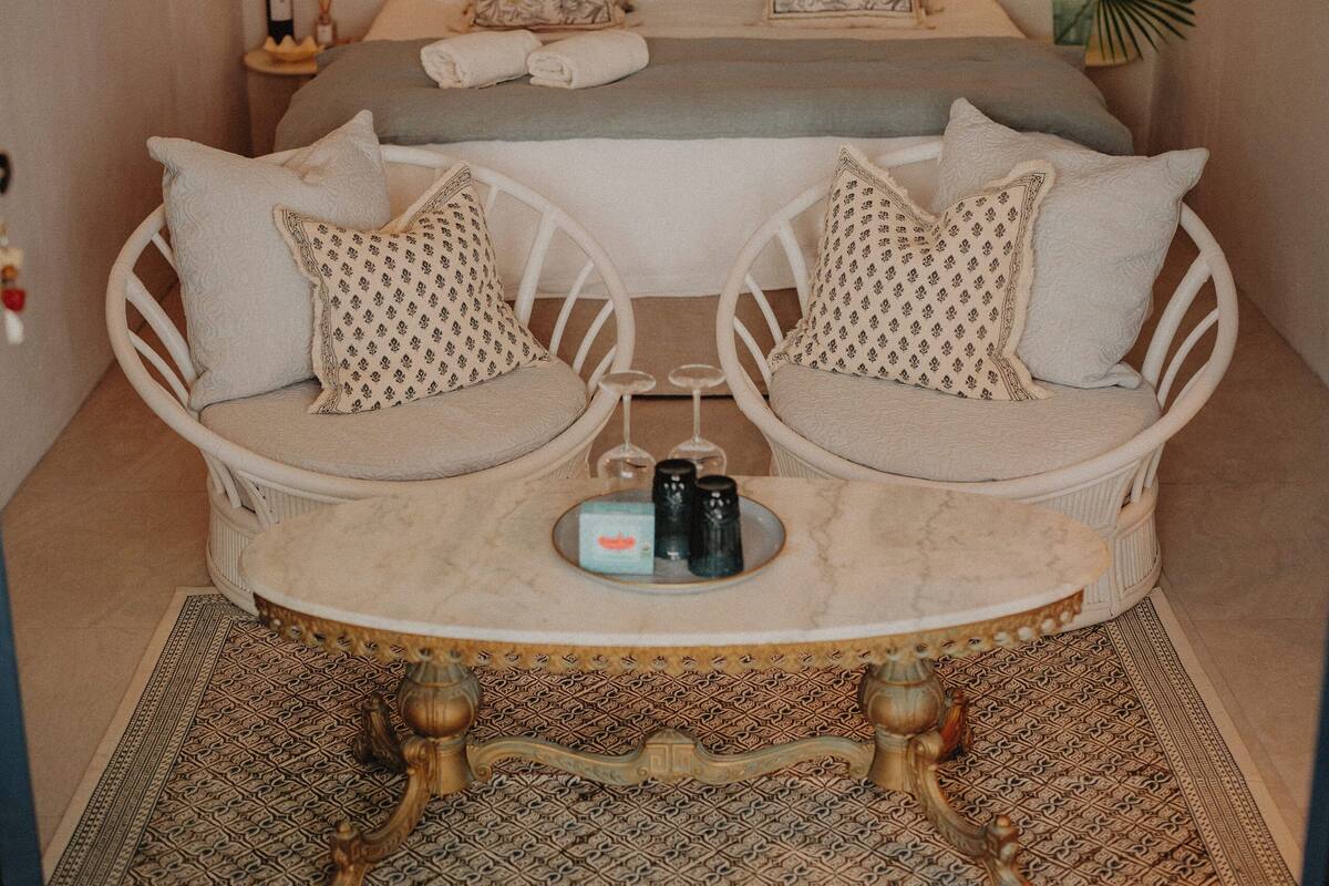 A bedroom with two white wicker chairs, a small round table, and a bed with multiple pillows.