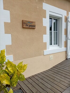 Ein Gebäude mit einer strukturierten, beige farbenen Wand, einem Holzschild mit der Aufschrift 'Le guêpier', einem Fenster mit einem gemusterten Rollo und einer Holzterrasse.