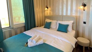 A hotel room with a large bed, two wall-mounted lamps, a window with curtains, and a small bedside table. - Sveti Stefan Panorama View Apartment (Sveti Stefan)