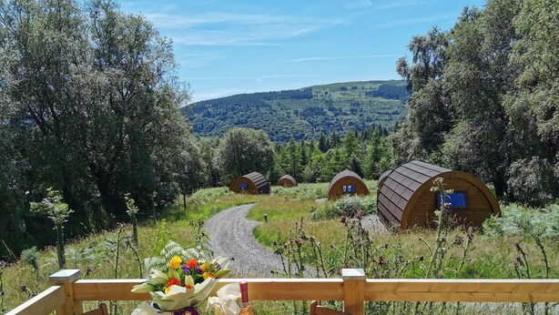 A wooden deck with a table, chairs, and a bottle of champagne. In the background, there are several wooden cabins surrounded by trees and a winding path. - Glamping Pod - Glendalough (Laragh)