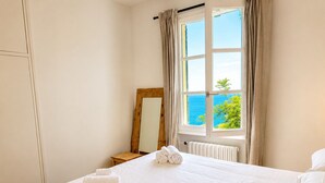 Ein Hotelzimmer mit Bett, einem Holzstuhl, einem Spiegel und einem Fenster mit Meerblick.
