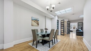 A dining area with a glass table and chairs, a chandelier, a painting, and a view through a door. - The Skylight Vault - Bright Downtown Loft Living/Pet Friendly/Short Walk to Shop (Sarnia)