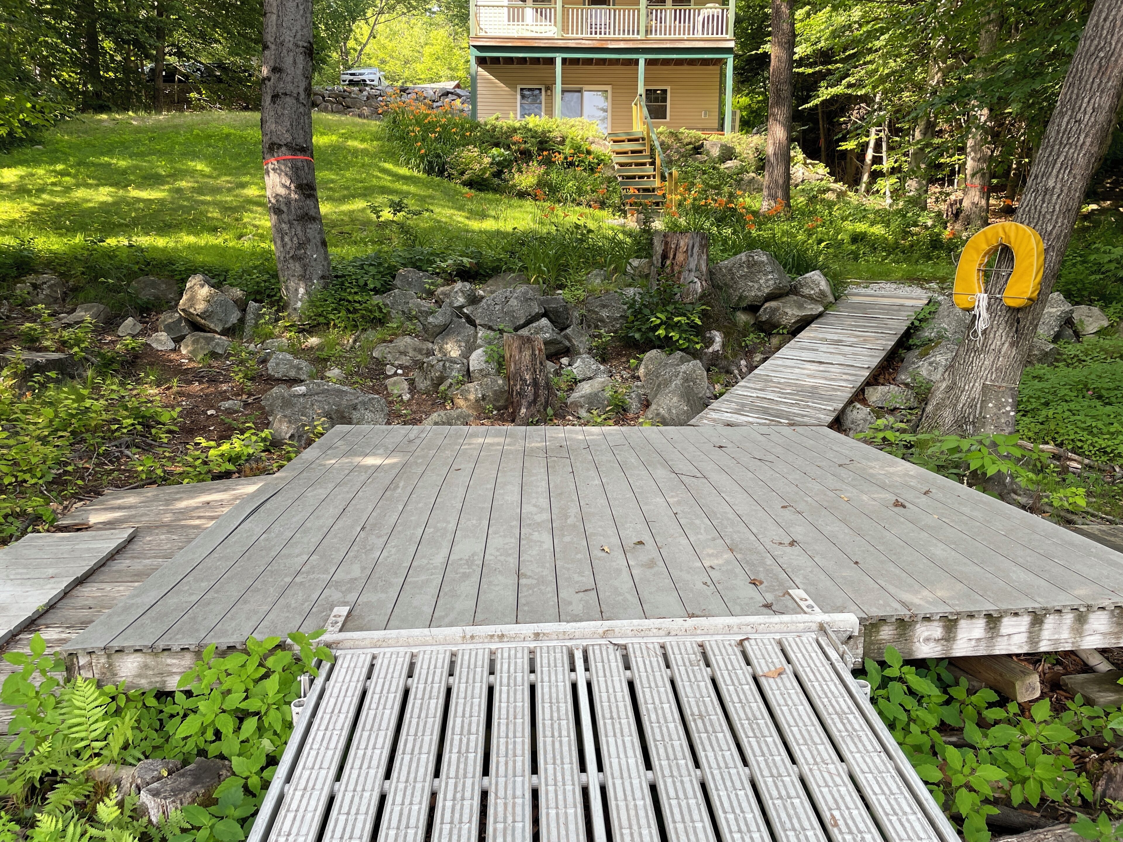 Une terrasse en bois menant à une maison dotée d’un balcon, le tout entouré de verdure et de rochers.