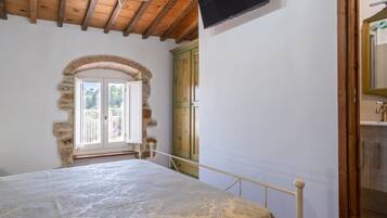 A bedroom with a bed, wooden ceiling beams, a stone archway, a wooden wardrobe, and a television mounted on the wall.