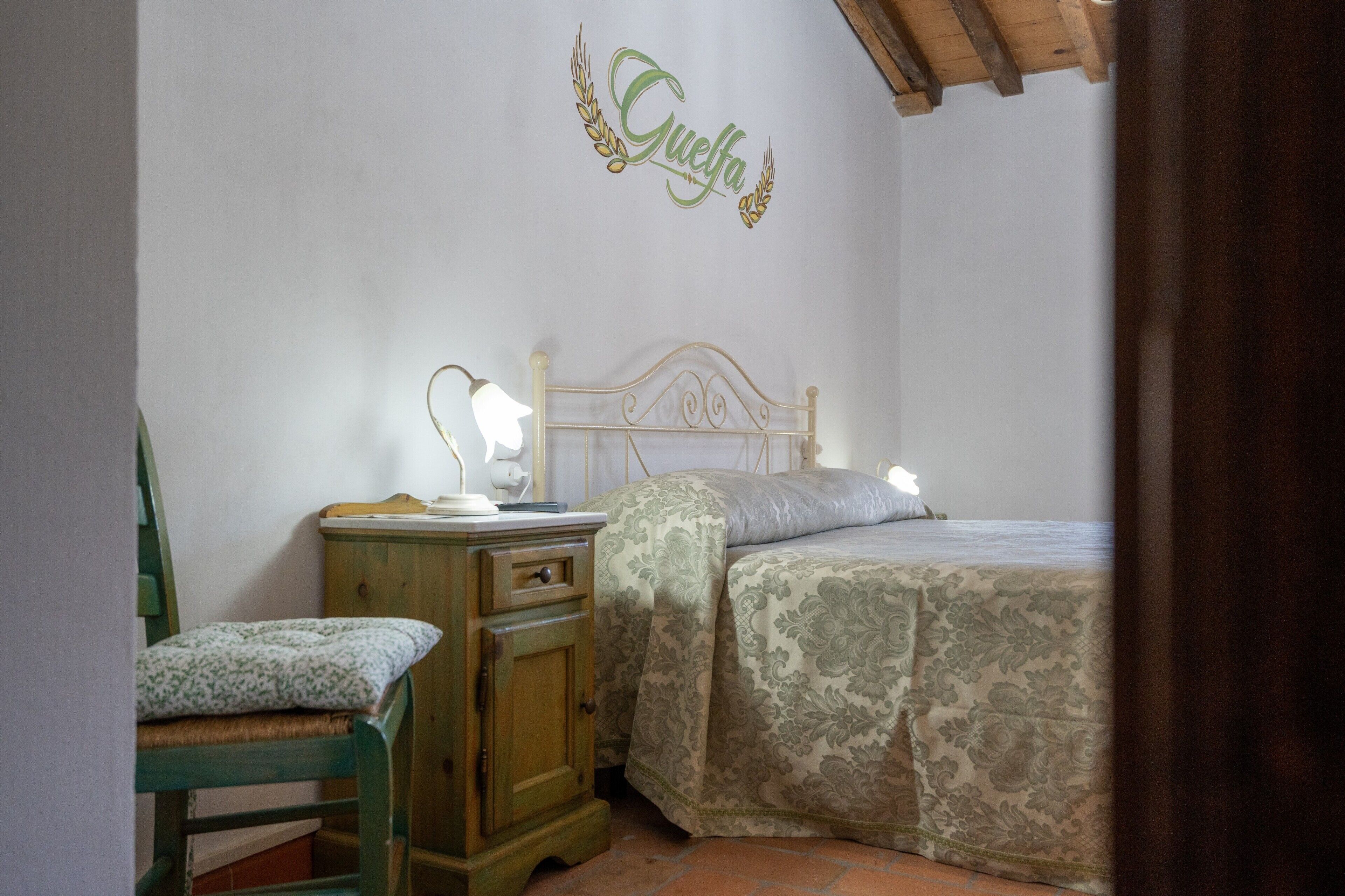 Un dormitori amb un llit, una tauleta de nit amb una làmpada, una cadira i una paret amb una decoració amb un nom.