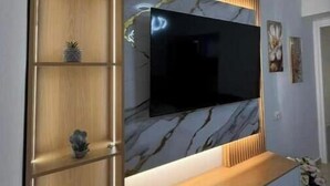 Ein modernes Wohnzimmer mit einem großen Flachbildfernseher, der an einer Wand mit marmorartigem Dekor befestigt ist. Holzregale mit dekorativen Gegenständen und ein Holzkonsolentisch befinden sich unter dem Fernseher.