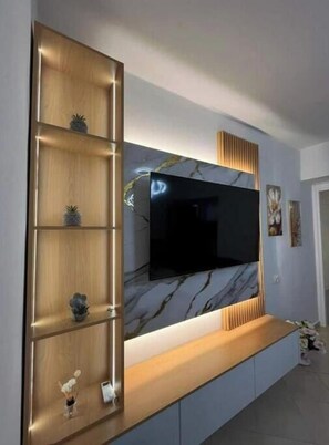 Ein modernes Wohnzimmer mit einem großen Flachbildfernseher, der an einer Wand mit marmorartigem Dekor befestigt ist. Holzregale mit dekorativen Gegenständen und ein Holzkonsolentisch befinden sich unter dem Fernseher.