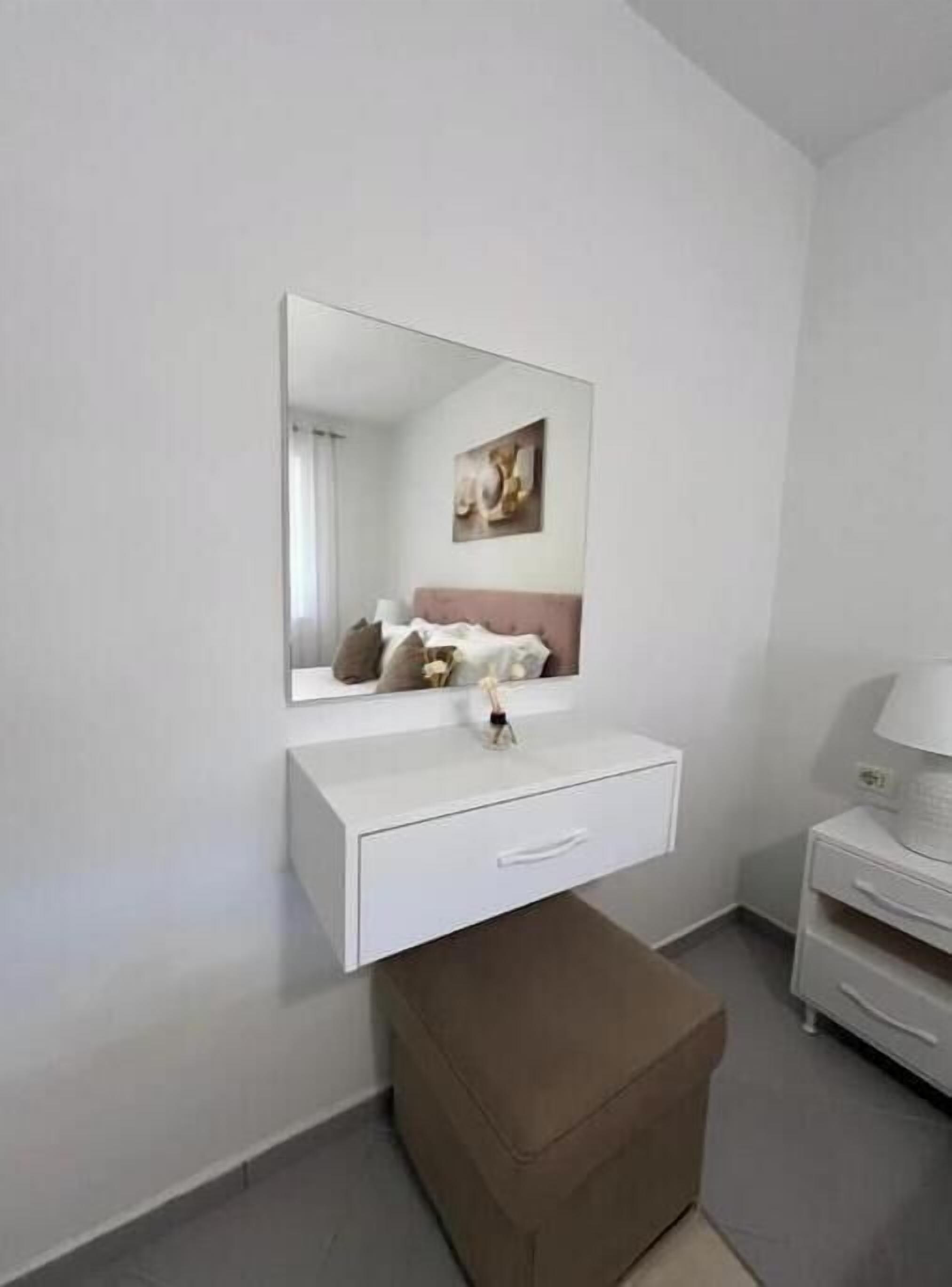 A white bedside table with a lamp, a mirror above it, and a brown ottoman in front.