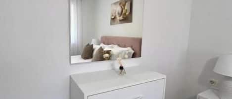 A white bedside table with a lamp, a mirror above it, and a brown ottoman in front.
