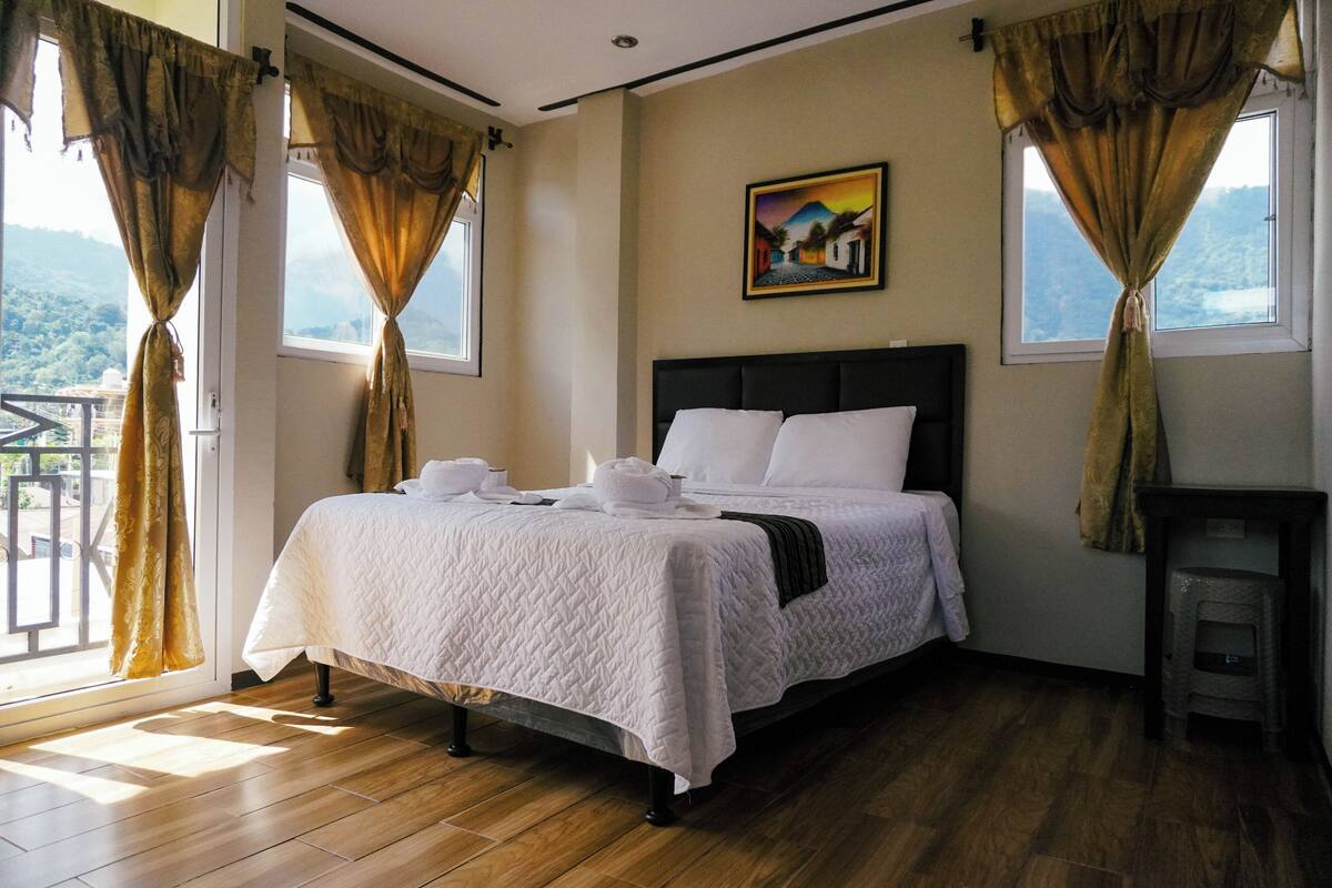 A neatly made bed with white linens, a painting on the wall, and a view through the window.