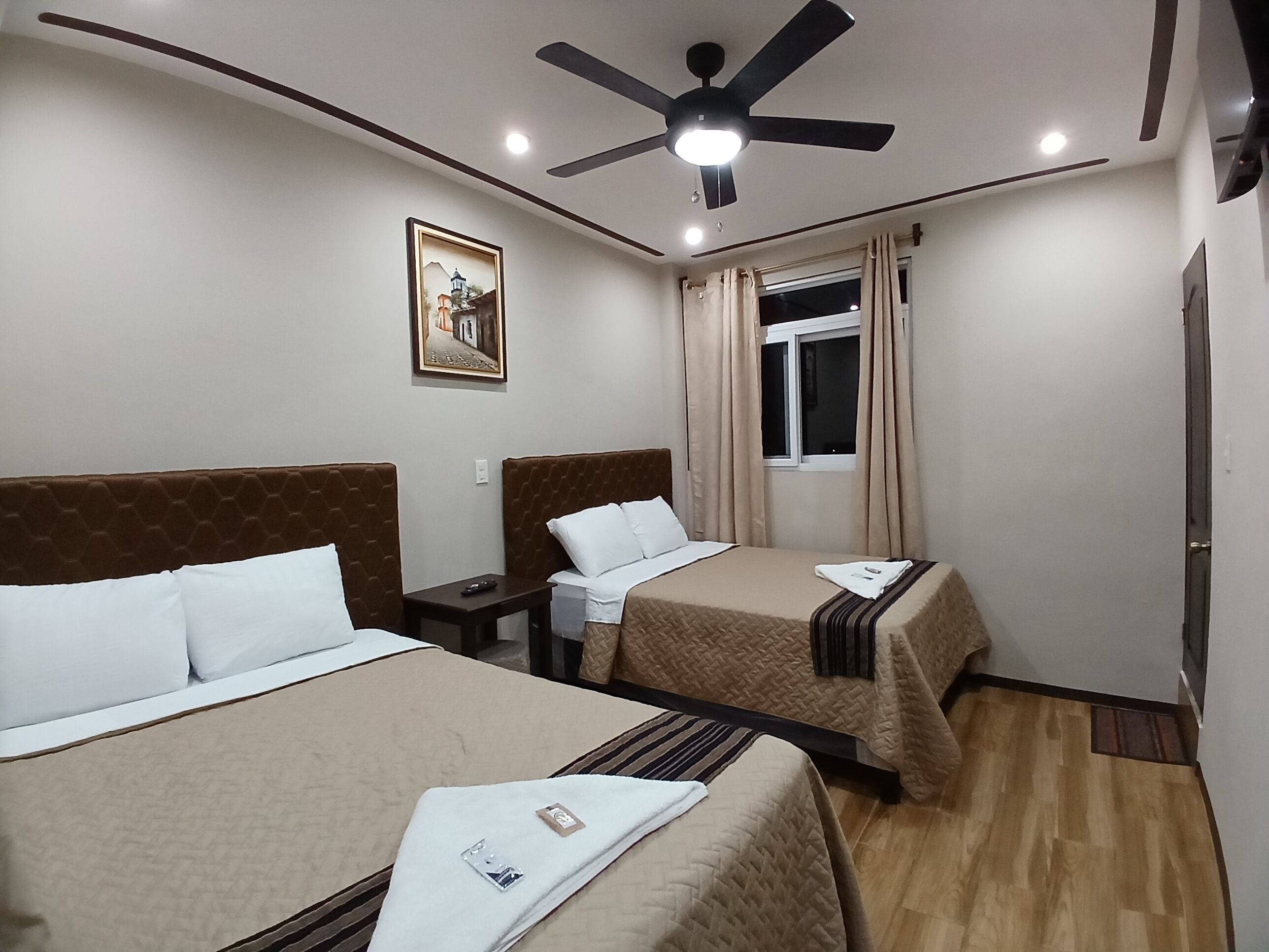 A hotel room with two beds, a ceiling fan, a window with curtains, a painting on the wall, and a nightstand with a remote control.