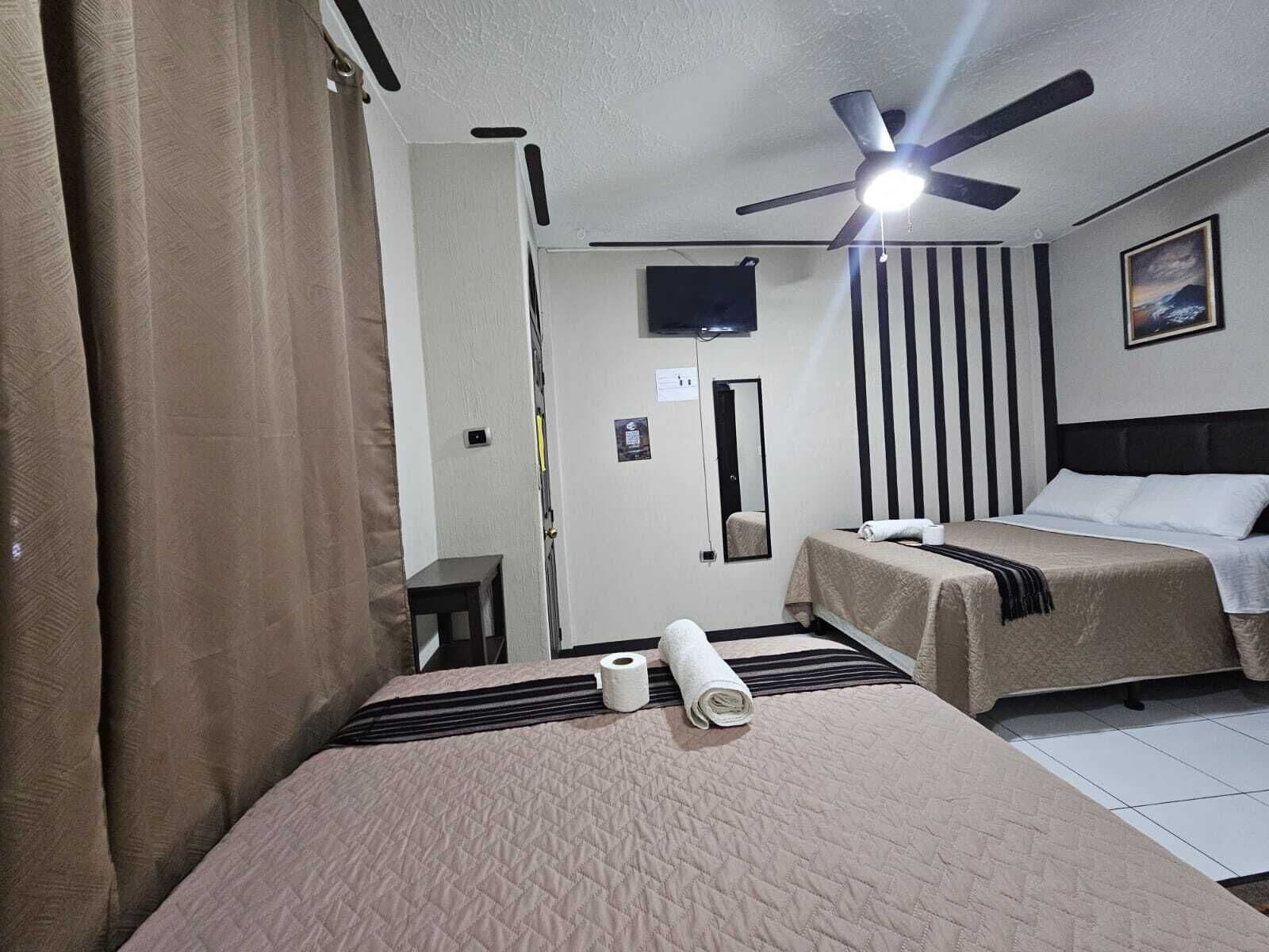 Hotellihuone, jossa on sänky, kattotuuletin, televisio, peili ja taulu seinällä.