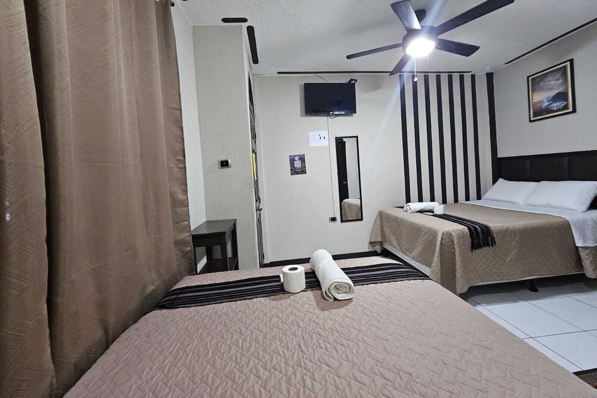 A hotel room with a bed, a ceiling fan, a TV, a mirror, and a painting on the wall.