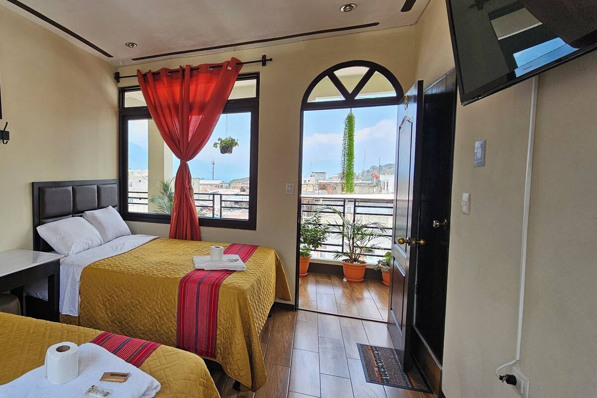 A hotel room with two beds, a desk, a window with red curtains, and a balcony with potted plants.