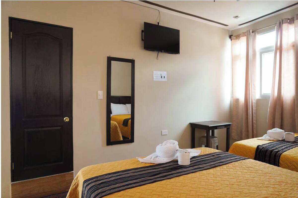 A hotel room with two beds, a TV, a mirror, a small table, and a window with curtains.