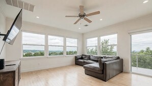 A spacious living room with a large window, a ceiling fan, and a leather sofa. - Dripping Springs Hill Country Retreat - Stunning Views + Game Room on 5 acres!! (Dripping Springs)