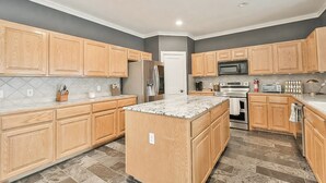 A modern kitchen with wooden cabinets, a granite countertop island, stainless steel appliances, and a tiled floor. - Dripping Springs Hill Country Retreat - Stunning Views + Game Room on 5 acres!! (Dripping Springs)