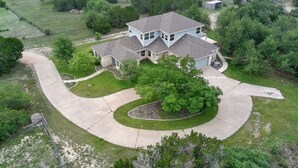 An aerial view of a two-story house with a large driveway, a tree in the front yard, and a fenced property. - Dripping Springs Hill Country Retreat - Stunning Views + Game Room on 5 acres!! (Dripping Springs)