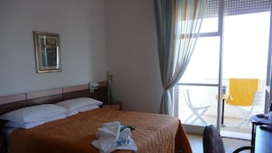 Una habitación de hotel con cama, una silla, una mesa, un ventanal con cortinas y una planta.