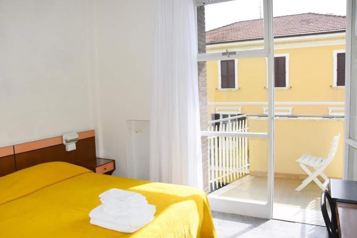 A hotel room with a bed, a nightstand, a window with curtains, and a balcony with a chair.