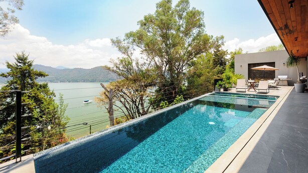 Piscina con vistas a un lago y montañas, mobiliario exterior y un edificio moderno con techo de madera.