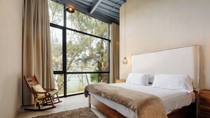 Un dormitorio con un ventanal grande, una mecedora de madera, una cama con sábanas blancas y una mesita de noche de madera.