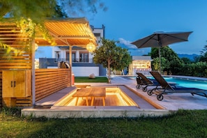 A poolside area with wooden deck seating, a pergola with string lights, and a view of a building with a bar. - The Infinities - Premium Villa Alexandros (Kos)