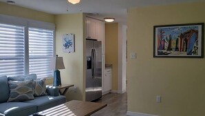Ein Wohnzimmer mit einer Couch, einem Tisch, einer Lampe, einem Kühlschrank und einem Bild an der Wand.