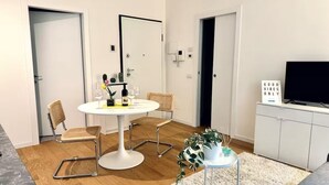 Una sala de estar moderna con una mesa de comedor redonda, dos sillas, un televisor de pantalla plana y una pequeña mesita auxiliar con una planta.