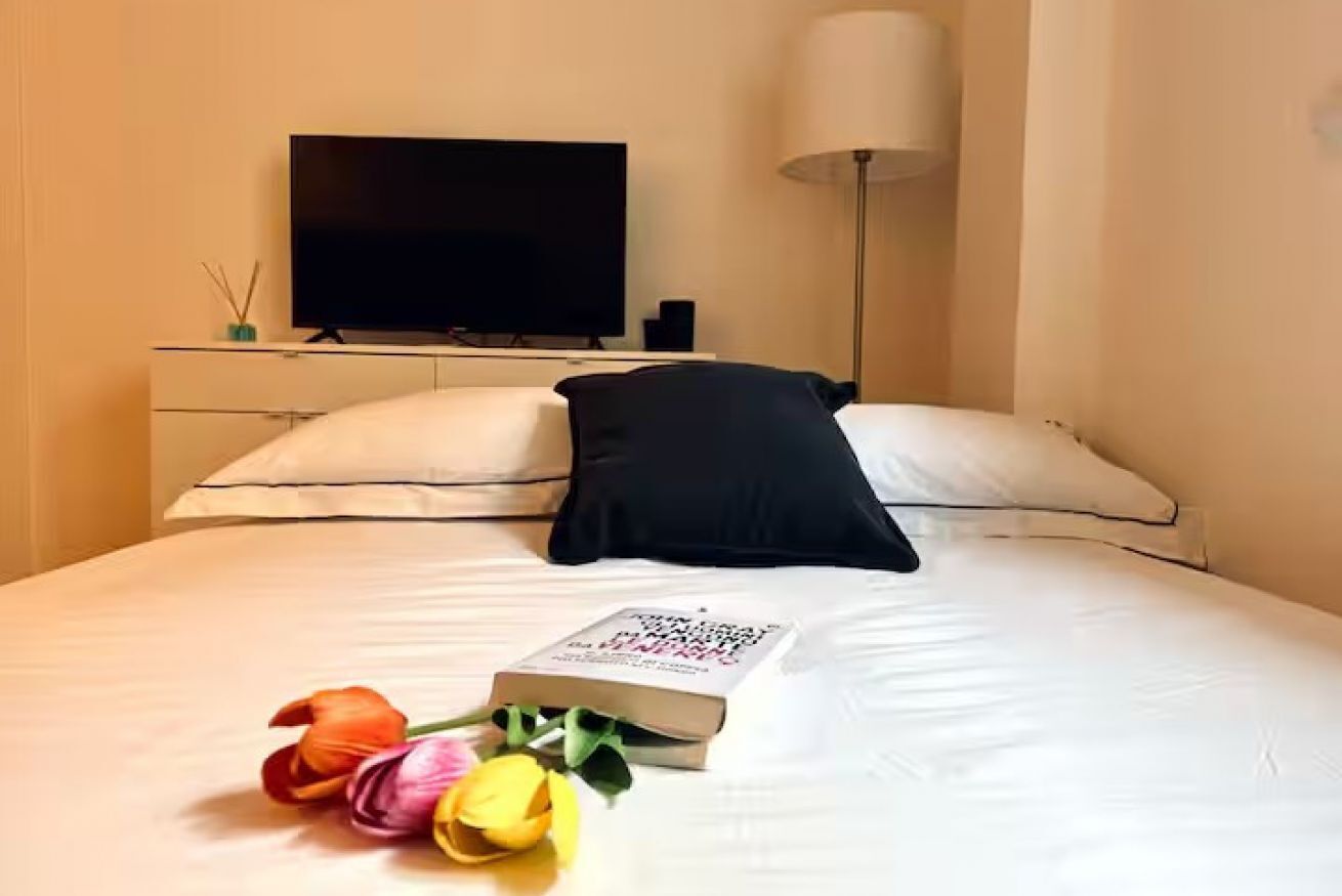 Ein Hotelzimmer mit einem Bett, einem Flachbildfernseher, einer Nachttischlampe und einem Buch mit Blumenstrauß.