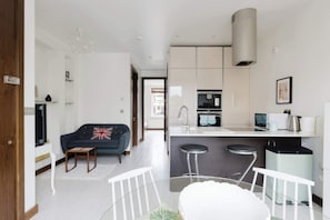 Eine moderne Küche mit Einbauherd, ein Essbereich mit Glastisch und ein Wohnzimmer mit Sofa und Fernseher.