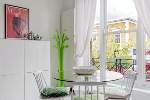 A dining area with a glass table, white chairs, a green decorative piece, and a window with a view of buildings and greenery. - Chic 2BDR East London Hideaway (Greater London)