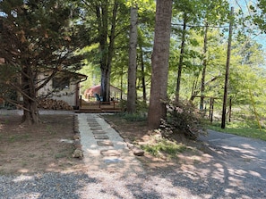 A rustic cabin with a gravel path leading to a wooden deck, surrounded by tall trees and a clear sky.