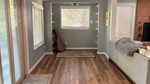 Una habitación con piso de madera, una guitarra apoyada en la pared, una ventana con persianas, un sofá con una manta y una chimenea.