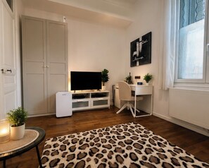 Ein kleines Zimmer mit Fernseher, Schreibtisch, Stuhl, Kleiderschrank und Fenster.
