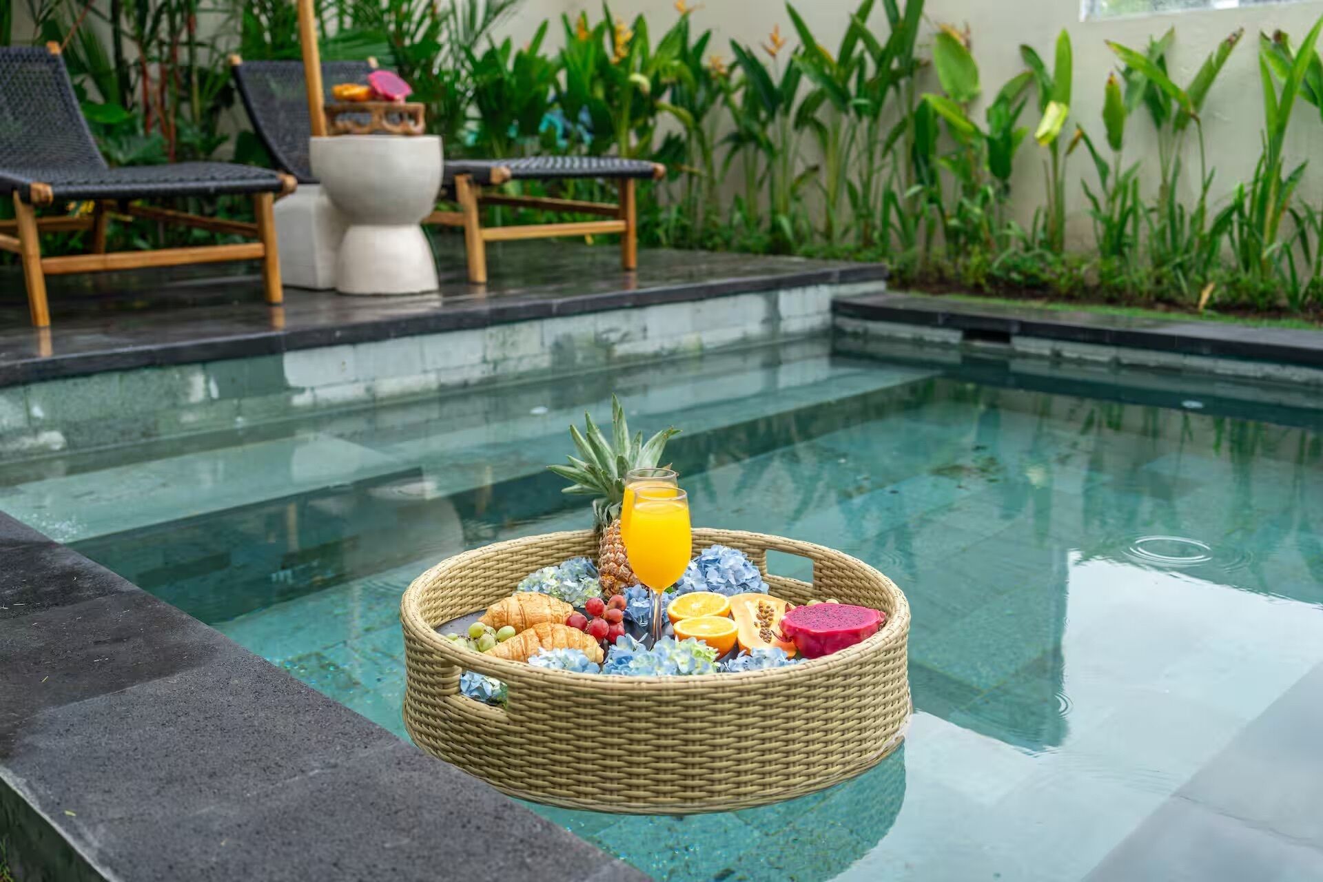 Ein Poolbereich mit einem Korbwicker-Korb, der Früchte und eine Ananas enthält, umgeben von Liegestühlen und einer tropischen Bepflanzung im Hintergrund.